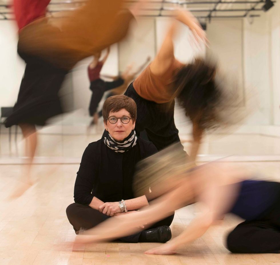 Gina Gibney sits on the floor of a dance studio, staring down the camera, as blurry dancers swirl around her
