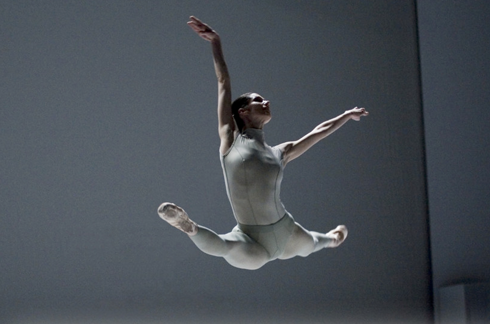 Wearing a grey unitard and against a grey set, Juleen leaps in an expansive grande jete, one arm extended above her and the other to the side