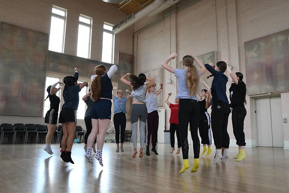 A group of teen girls jump in a large studio jump in the air, facing each other in a circle.