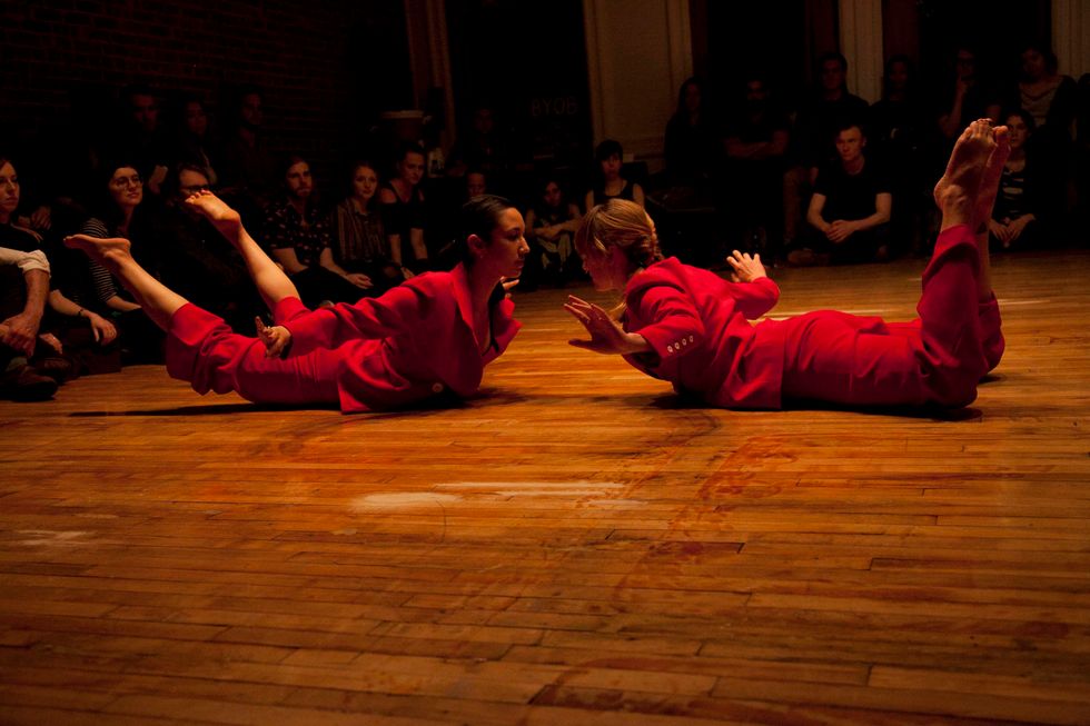 Erica Saucedo and another dancer lie on the ground on their stomachs facing each other, holding their arms, chest and legs off the ground. They wear red pant suits, and the audience sits around them.