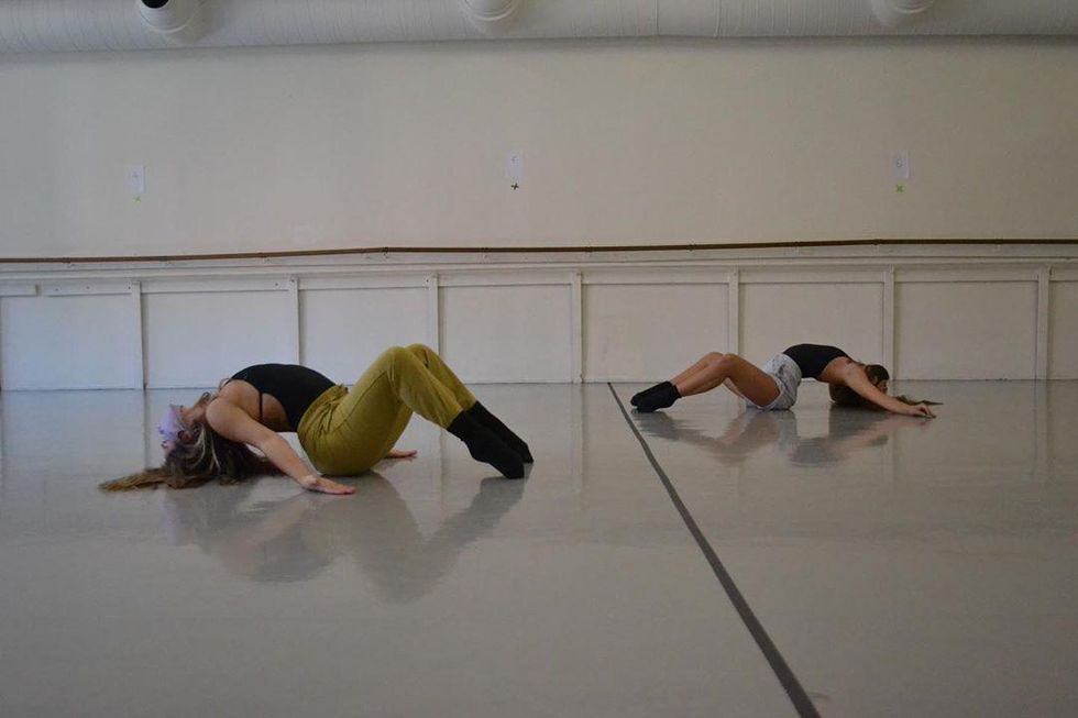 Two students wearing masks mirror each other on either side of a taped dividing line running down the middle of the studio. They both sit on the ground, arching back so the tops of their heads touch the floor, with their legs bent and toes pointed