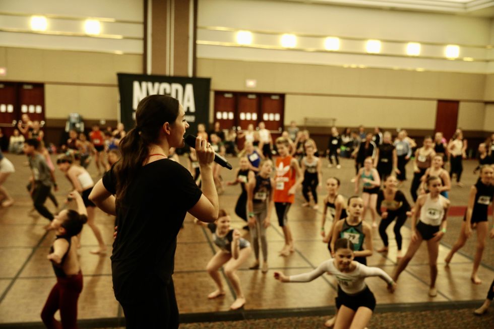 Grace Buckley stands on a convention stage, speaking into a microphone. Young dancers dance in front of her on the ballroom floor