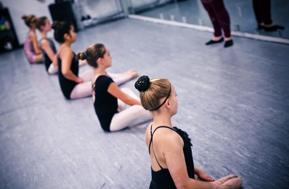 Five students sit on the floor of a dance studio in a straight line, wearing leotards and tights.