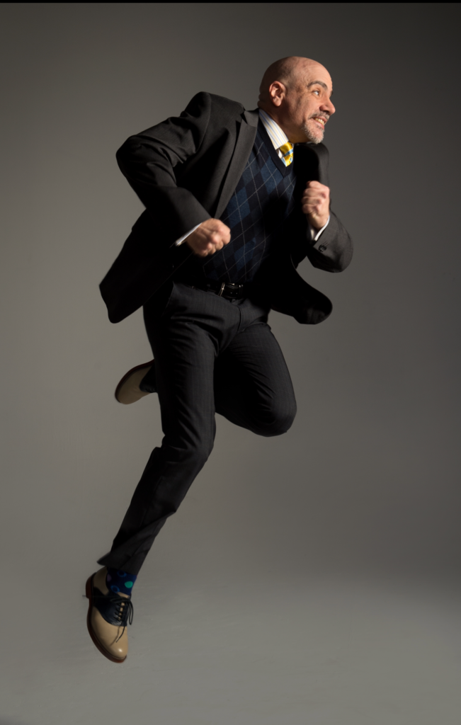 Bill Waldinger, a bald white man, wears a suit with a checkered sweater underneath. He jumps in the air in a running position, tucking one leg under him