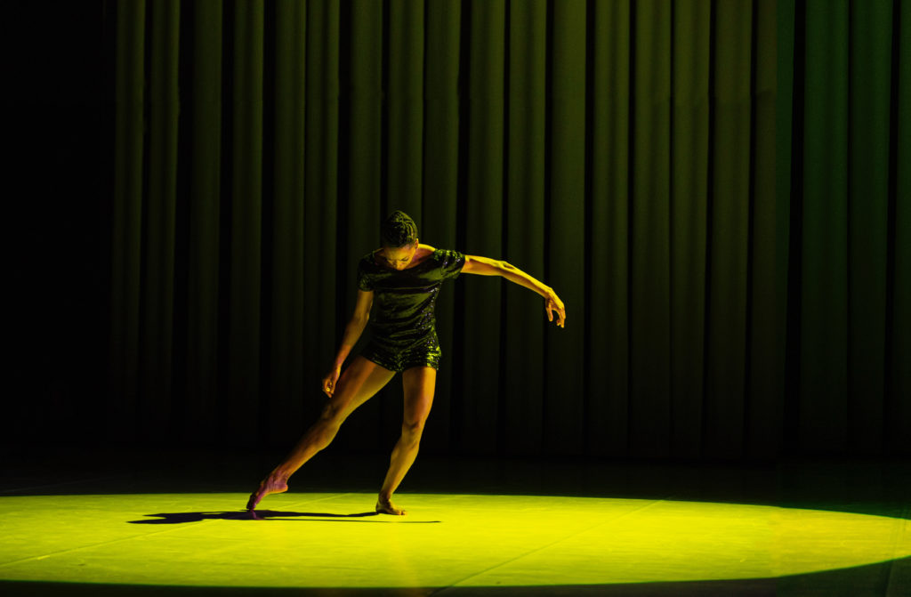 Tamisha Guy, a young Black woman wearing black shorts and a black shirt, dances in a pool of yellow light, in front of a black curtain. She tendus one leg to the side and looks down, extending her other arm to the other side.