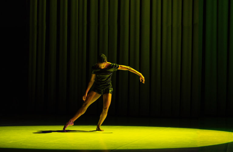 Tamisha Guy, a young Black woman wearing black shorts and a black shirt, dances in a pool of yellow light, in front of a black curtain. She tendus one leg to the side and looks down, extending her other arm to the other side.