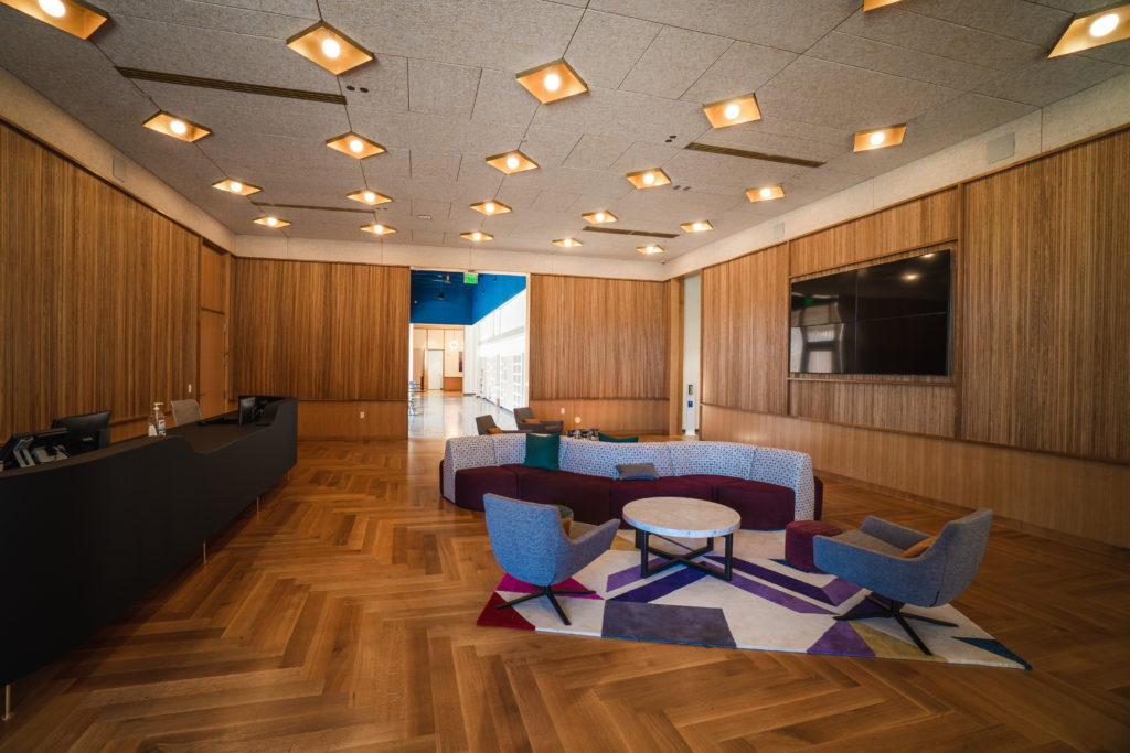 The lobby of Collage's new building, with colorful, trendy furniture, sleek overhead lighting, a front desk, a tv and wood paneled walls.