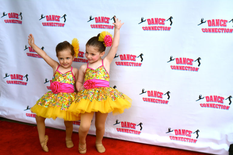 Two preschool-aged girls in sparkly yellow and pink costumes pose together on a red carpet. A backdrop with the Dance Connection logo is behind them