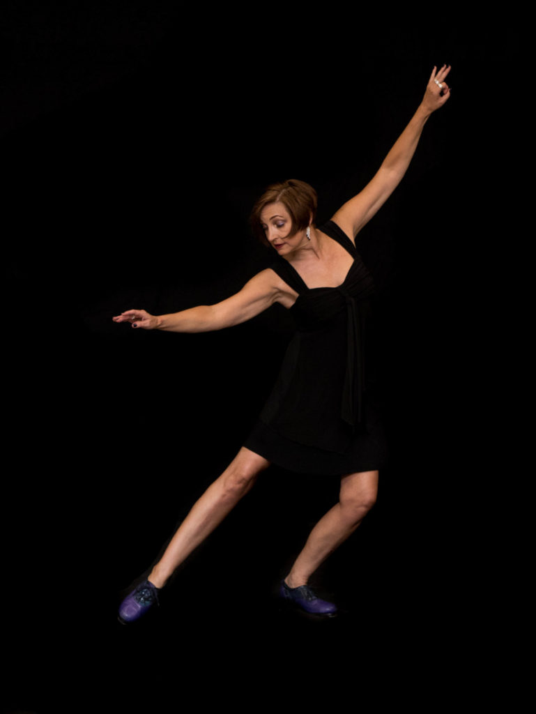 Cheryl Simmons, a white woman with short brown hair, wears a black dress and purple tap shoes. She stretches one leg out to the side and leans over, looking at her foot