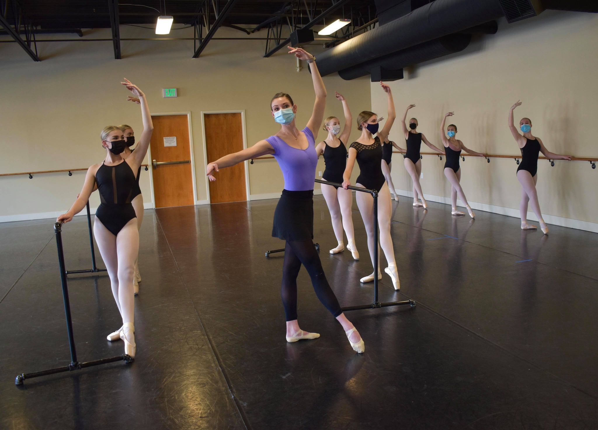 Danielle Dreis, dressed in a lavender leotard and black ballet skirt and tights with mask on, leads class in the front of a studio, positioned in Croisé devant. Other students in black leotards and pink tights and black masks copy her behind.