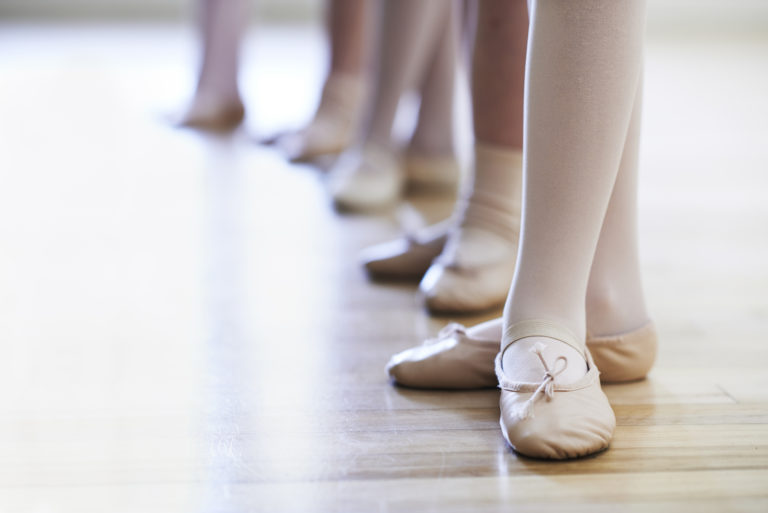 A close up of children's feet in ballet class. They wear pink tights and shoes, and are lined up in a row.