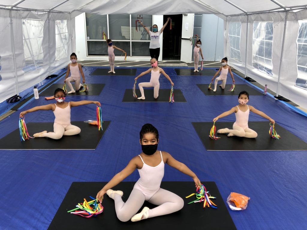 Nine young dancers and a teacher pose inside a tent in a parking lot. They hold rainbow ribbons and wear masks.