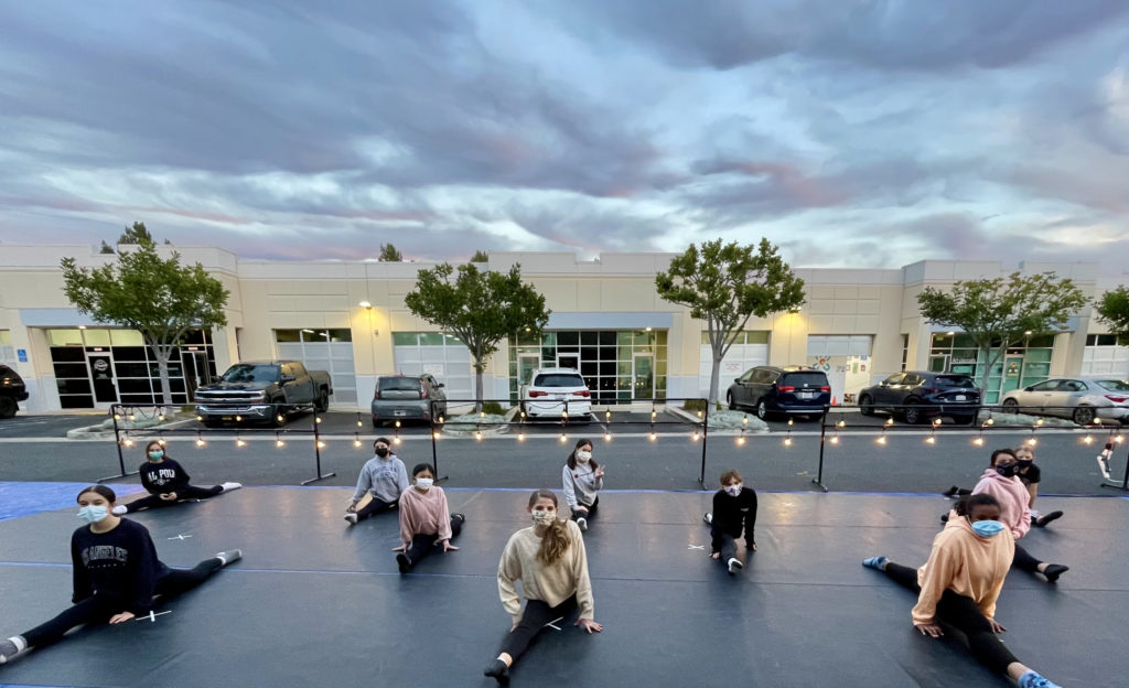 A group o teenage girls, all wearing masks, are in the splits on an outdoor stage in a parking lot. It is lit by fairy lights, and there is a business complex and some dramatic clouds behind them. 