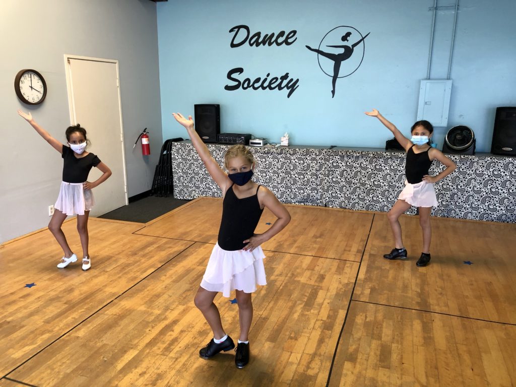 Three young dance students in masks pose in their dance studio, which has the Dance Society logo on the blue wall behind them. 