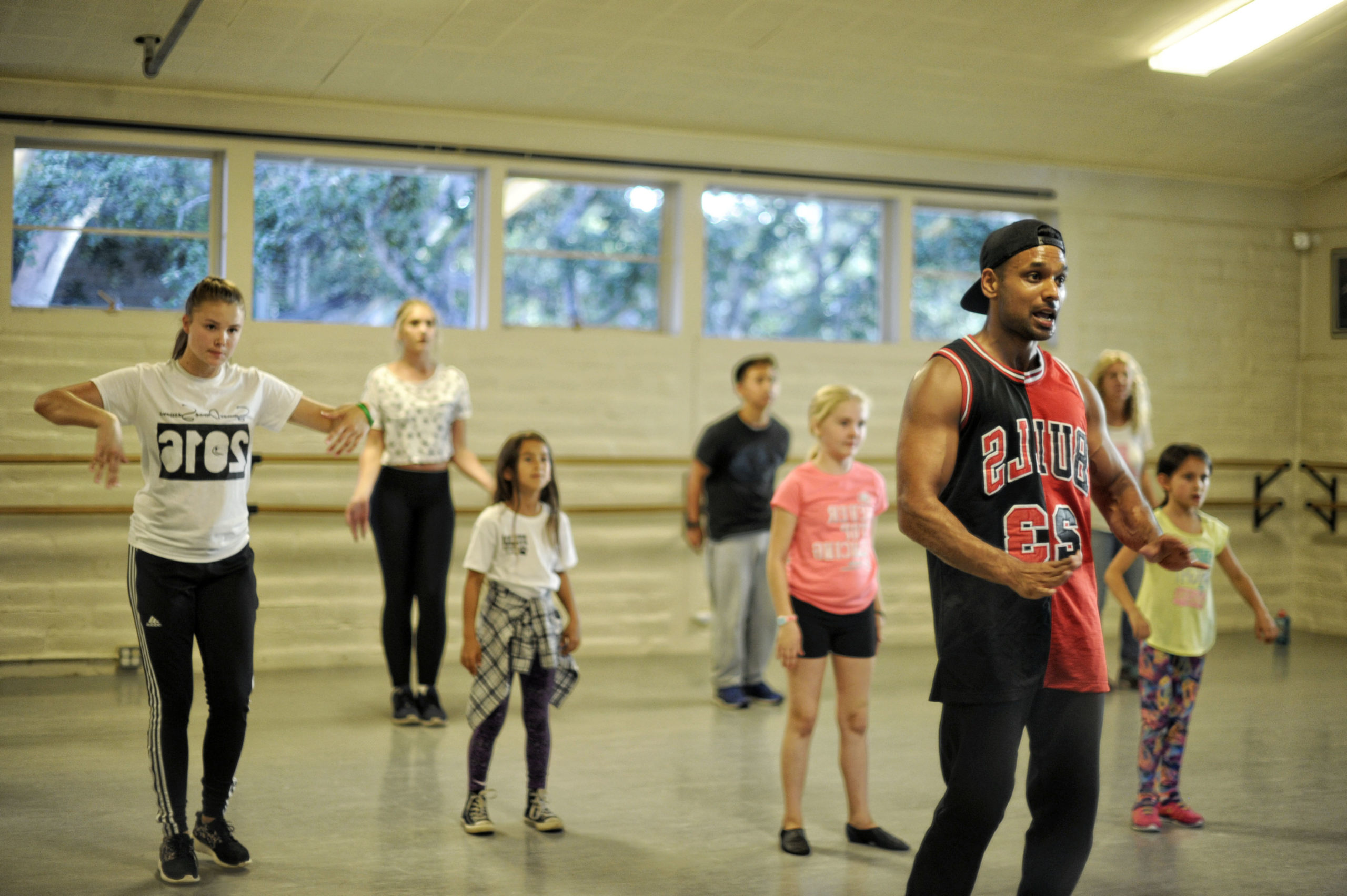 Pavan Thimmaiah, a young Indian-American man, teaches a dance class of a variety of ages. He wears a Bulls tank top and a backwards baseball cap, and students stand behind him in the studio.