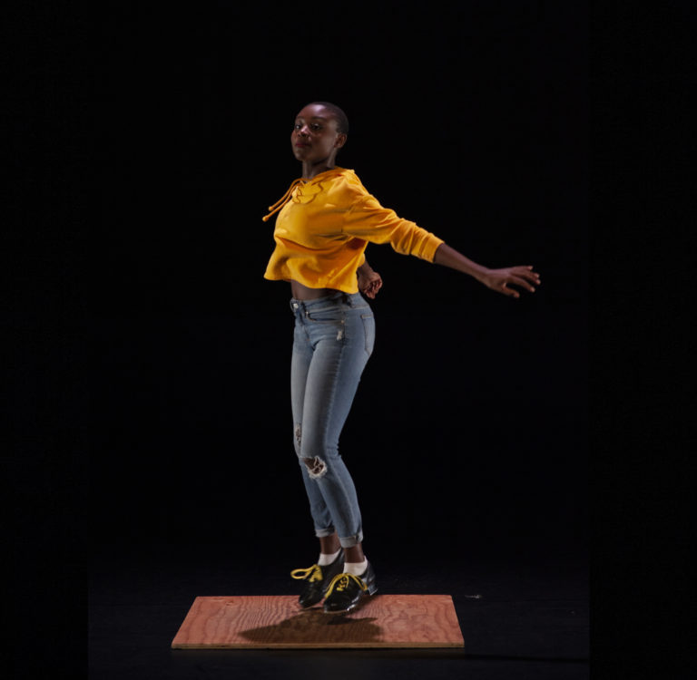 Micah Sallid, a young Black woman, dances on a small tap board. She wears jeans, an orange sweatshirt and black tap shoes.