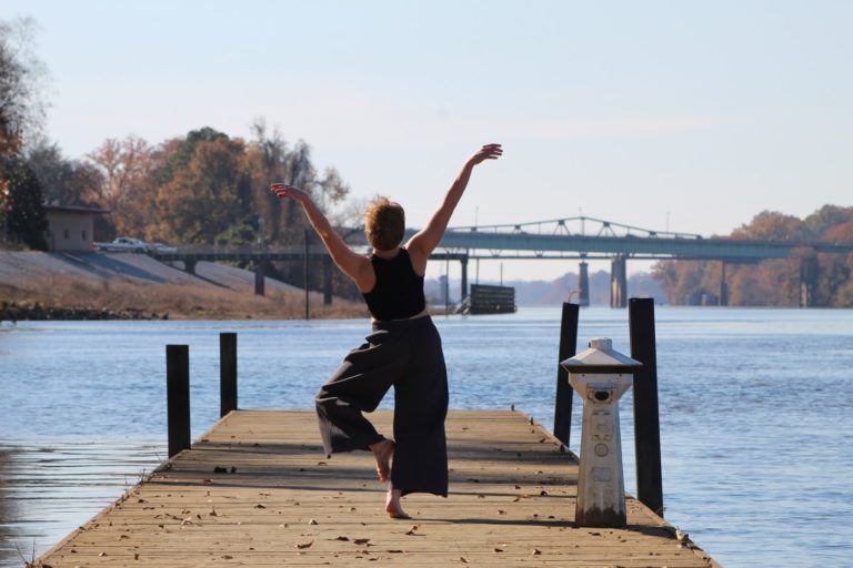 Facing away from the camera, Fen Kennedy dances on a dock, arms stretched upward and one leg bent behind them. Blue water, trees and a bridge are in the background.