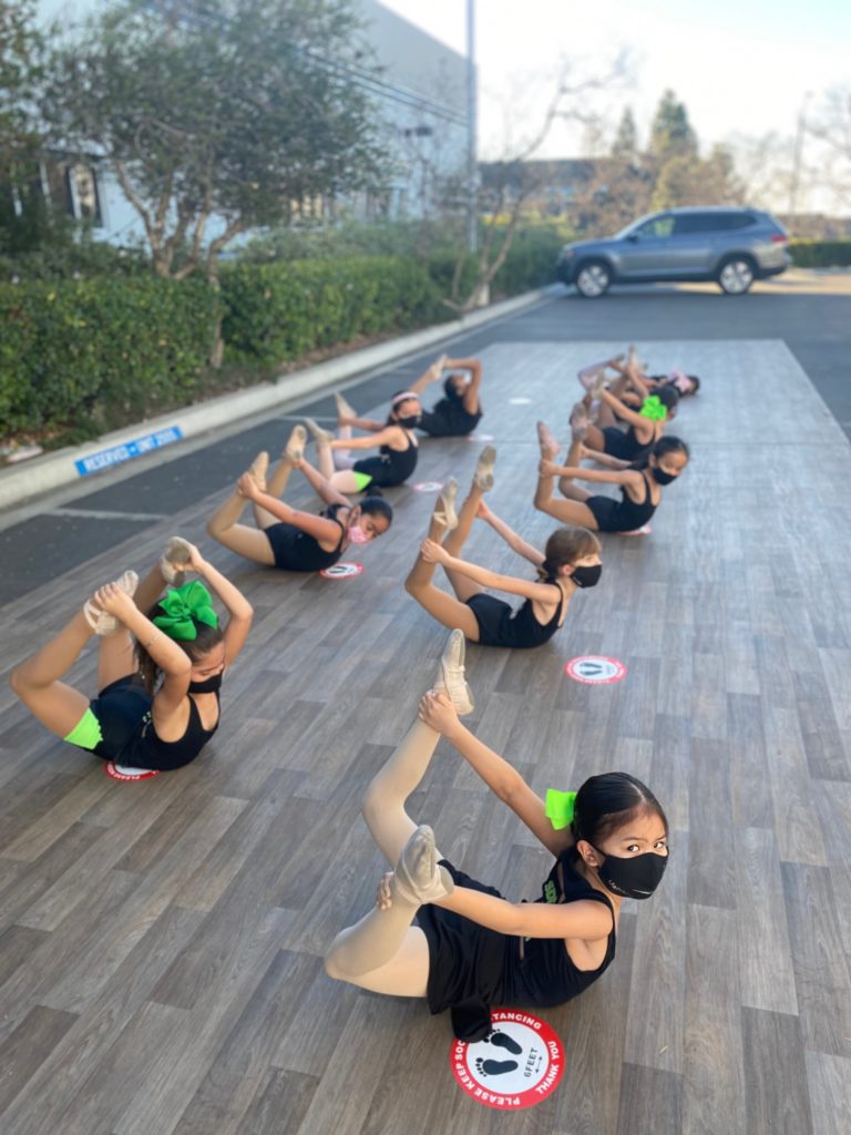 A group of young masked dancers stretch on an outdoor studio in a parking lot. They lay on their stomachs and reach behind them to grab their ankles.