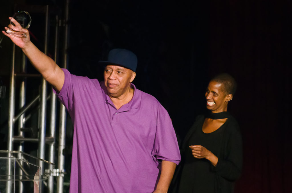 Frank Hatchett, an older Black man in a purple polo shirt, holds an award up in the air
