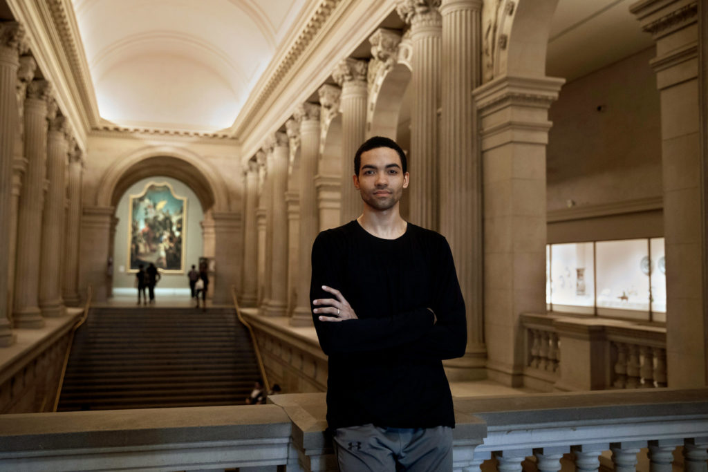 Silas Farley, a tall, slender, lighter-skinned Black man, leans against a stone balustrade in New York City's Metropolitan Museum of Art, arms crossed as he looks seriously at the camera.