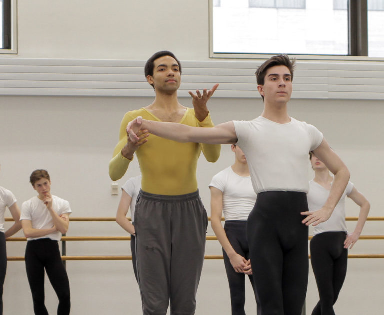 Silas Farley, a tall, slender Black man, here wearing grey sweat pants over a chartreuse biketard, gently lifts the arm of a male ballet student, making a 'lift up' gesture with his free hand.