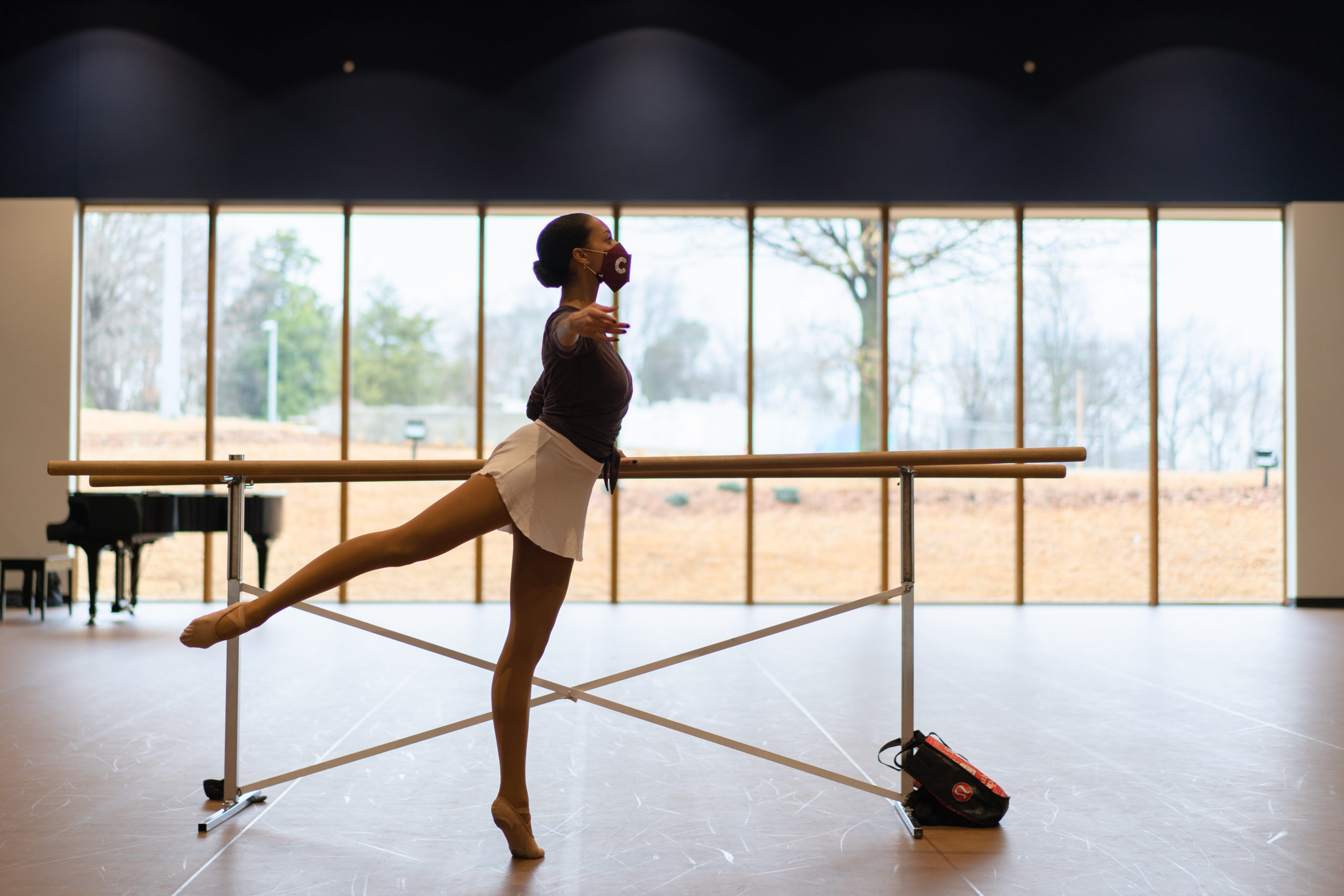 A dancer stands in releve arabesque at the barre, wearing a mask. She is in a large studio with a wall of windows behind her, overlooking some trees.
