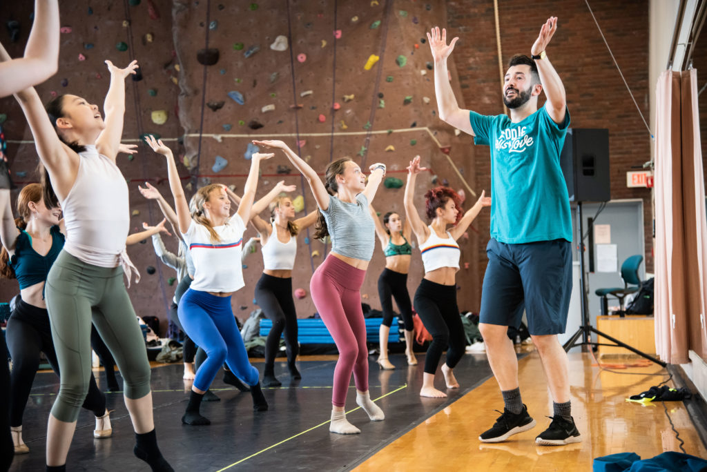 Al Blackstone, wearing a teal t-shirt and shorts, raises his hands in the air as if about to clap. A class full of young adult students face him and do the same thing.