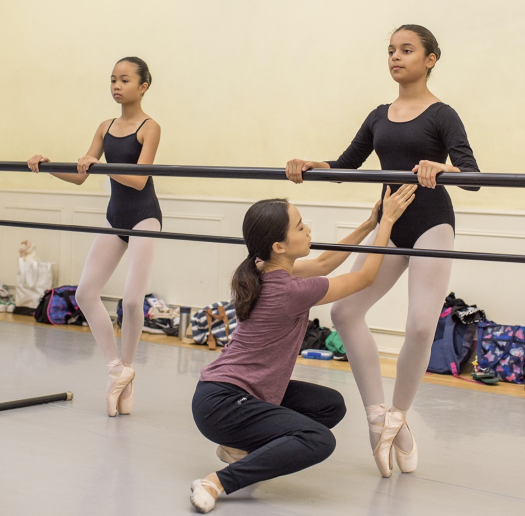 Alice Kim, wearing a purple shirt and black pants, kneels down to adjust a dancer's pelvic alignment. The dancer is at the barre, en pointe. 