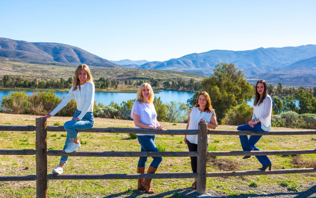 Neisha Hernandez, Kaylee Kiff, Franchesca Gonzalez Otañez and Melissa Treziok pose on a wooden fence overlooking beautiful lake and some mountains. They each wear white blouses and jeans.