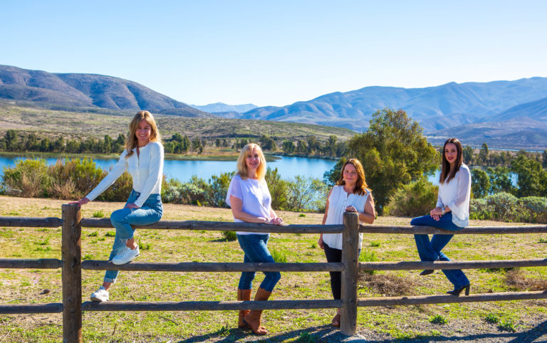 Neisha Hernandez, Kaylee Kiff, Franchesca Gonzalez Otañez and Melissa Treziok pose on a wooden fence overlooking beautiful lake and some mountains. They each wear white blouses and jeans.