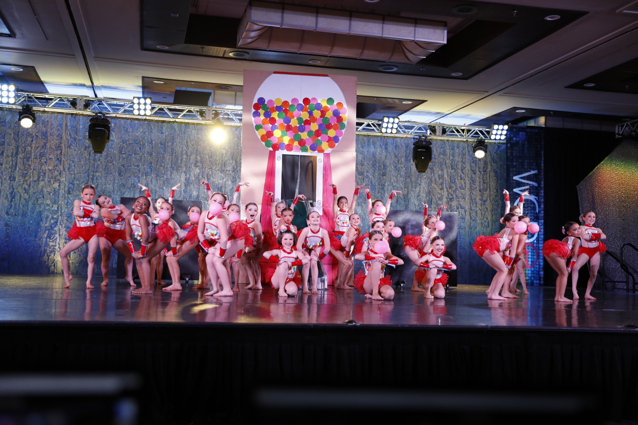 A large group of young dancers dance in a competition routine. Their costumes and backdrop are themed around a gumball machine.