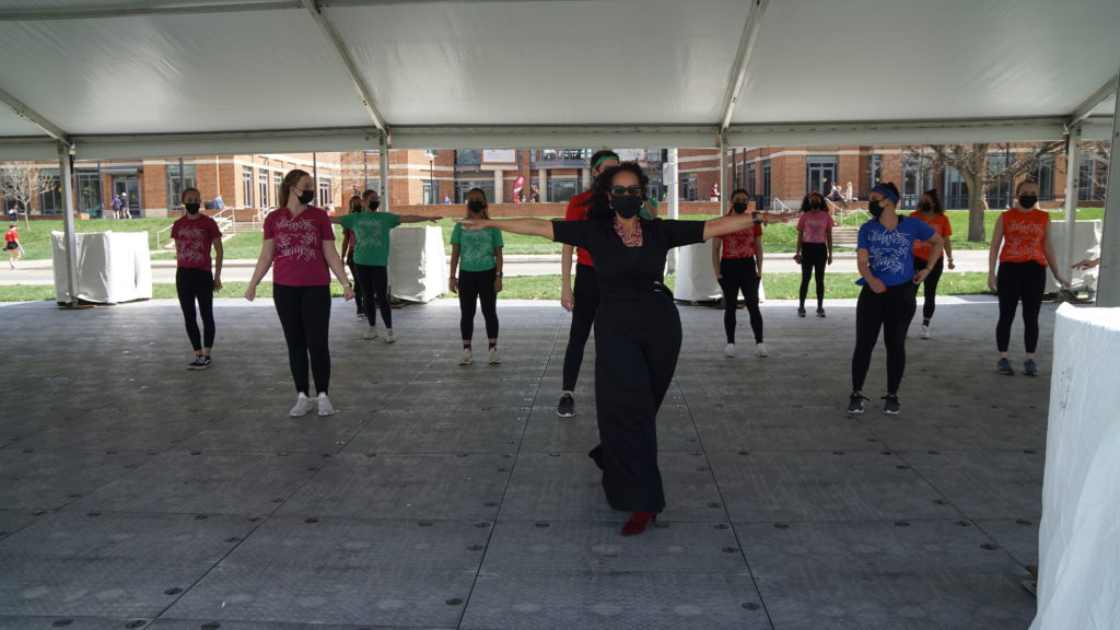 Dr. Nyama McCarthy Brown, a black woman wearing a mask, a chunky necklace and heels with her black outfit, leads an outdoor class, gesturing to the sides. A group of young women in t shirts, black pants and sneakers look on behind her.