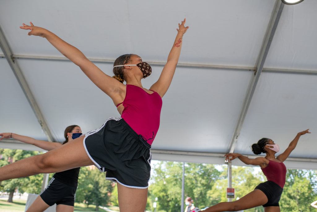 At an outdoor ballet class under a tent, student jump, with the arms in first position.