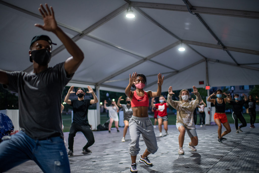 Students in sneakers and hip hop attire dance under an outdoor tent. With their heels off the ground, they turn slightly to the side with their palms up facing away from them.