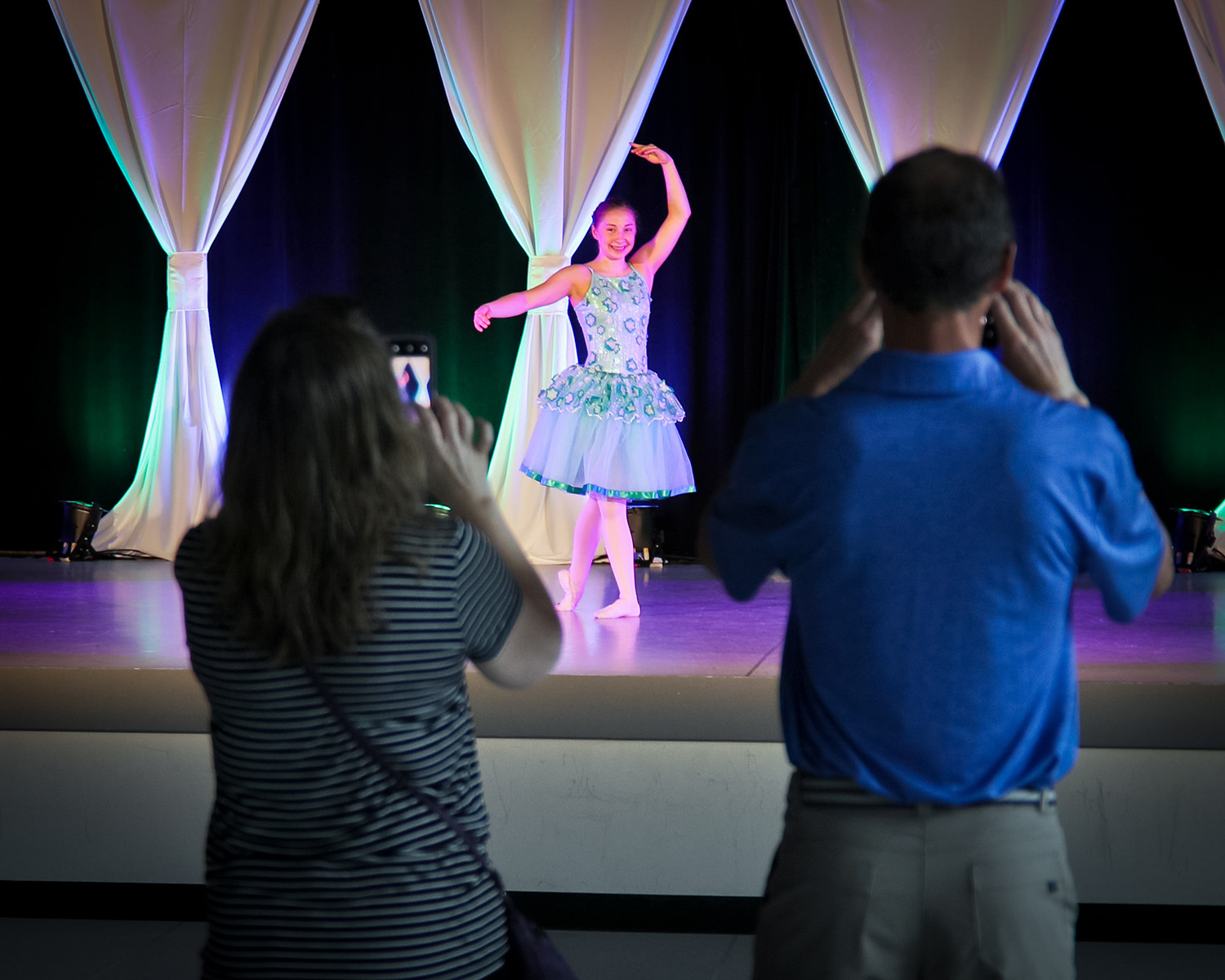 A middle school aged dancer performs a solo in a blue ballet costume with a long tutu. Two adults stand in front of the stage, taking photos or videos of her.