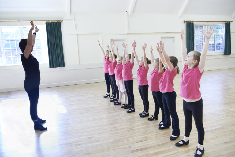 A line of girls in pink shirts and tap shoes raise their hands up to the sky, mimicking their teacher who stands in front of them.