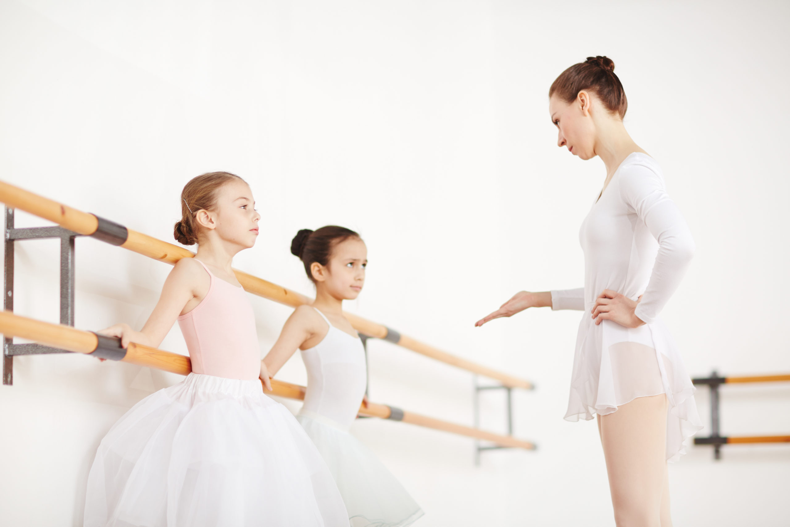 A ballet teacher in a white leotard and skirt talks sternly to two little ballet students during ballet class.