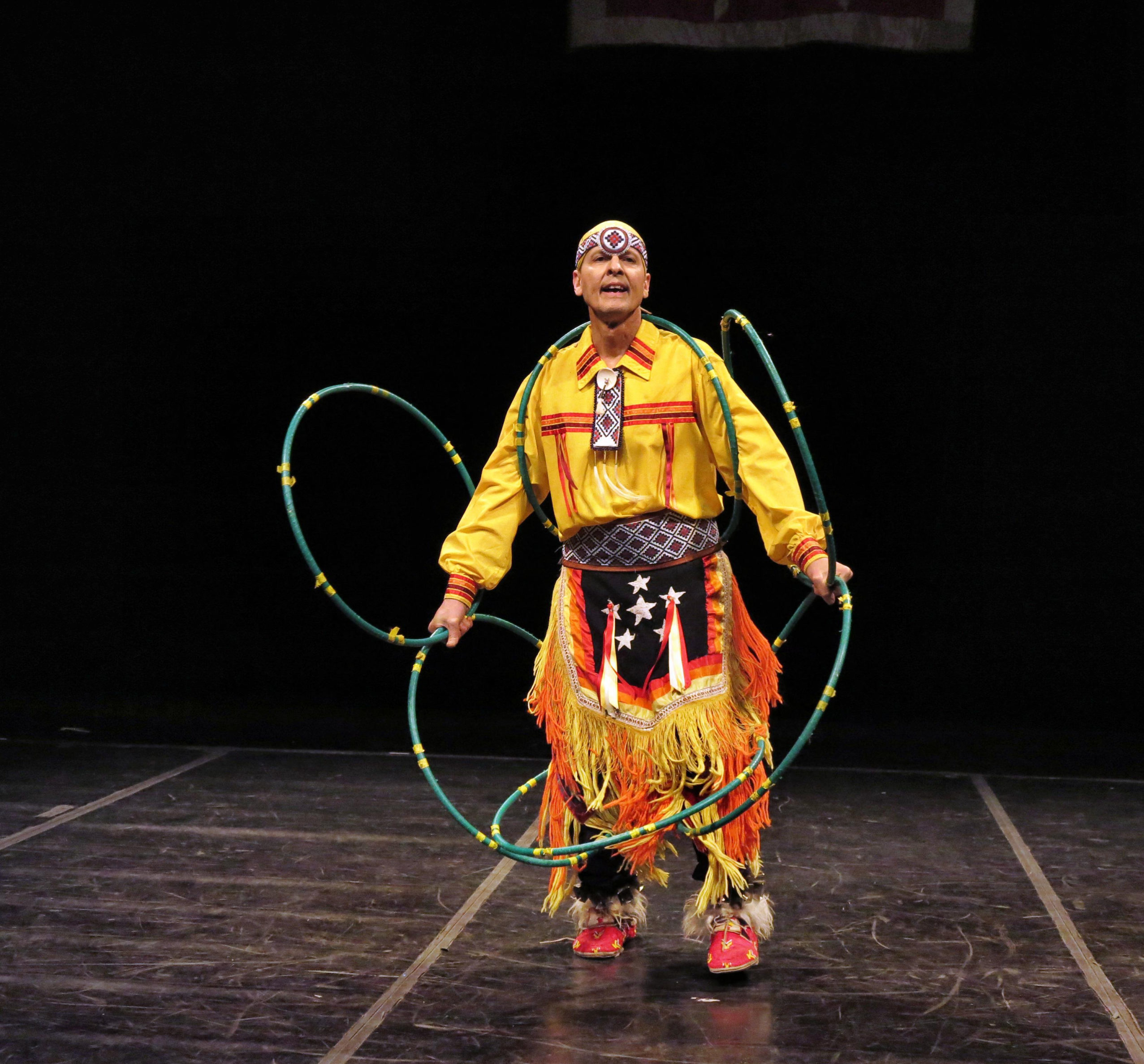 Michael Taylor, wearing a traditional yellow costume with red and black details, performs a hoop dance.
