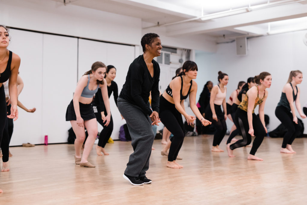 Sheila Barker, wearing dark workout clothes, stands in front of her class of jazz students and crouches slightly, leading them through a groovy movement while wearing a big smile.