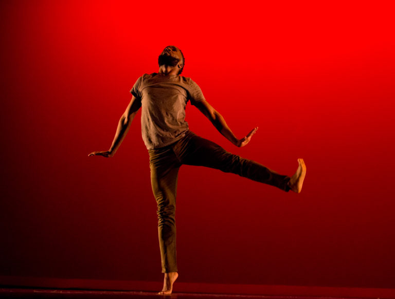 A young Black man, wearing a brown tshirt and darker brown pants, leaps against a red backdrop, flexing his feet and slightly arching his back, looking up.