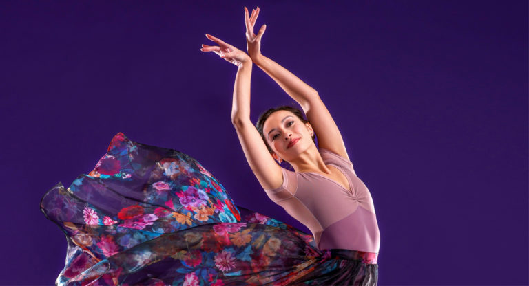Sarah-Gabrielle Ryan is shown from the waist up, smiling serenely to the camera and lifting her arms above her head with the back of her wrists touching together. She stands in front of a bright purple backdrop and wears a pink leotard and long, multi-colored floral skirt, which billows high behind her.
