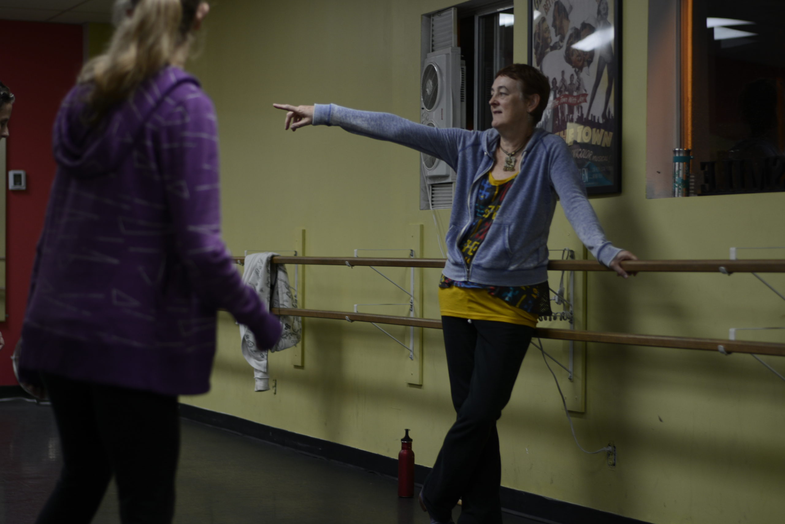 Heather Cornell, a white woman with short brown hair, leans against a ballet barre and points to the side, speaking to a woman who stands in front of her
