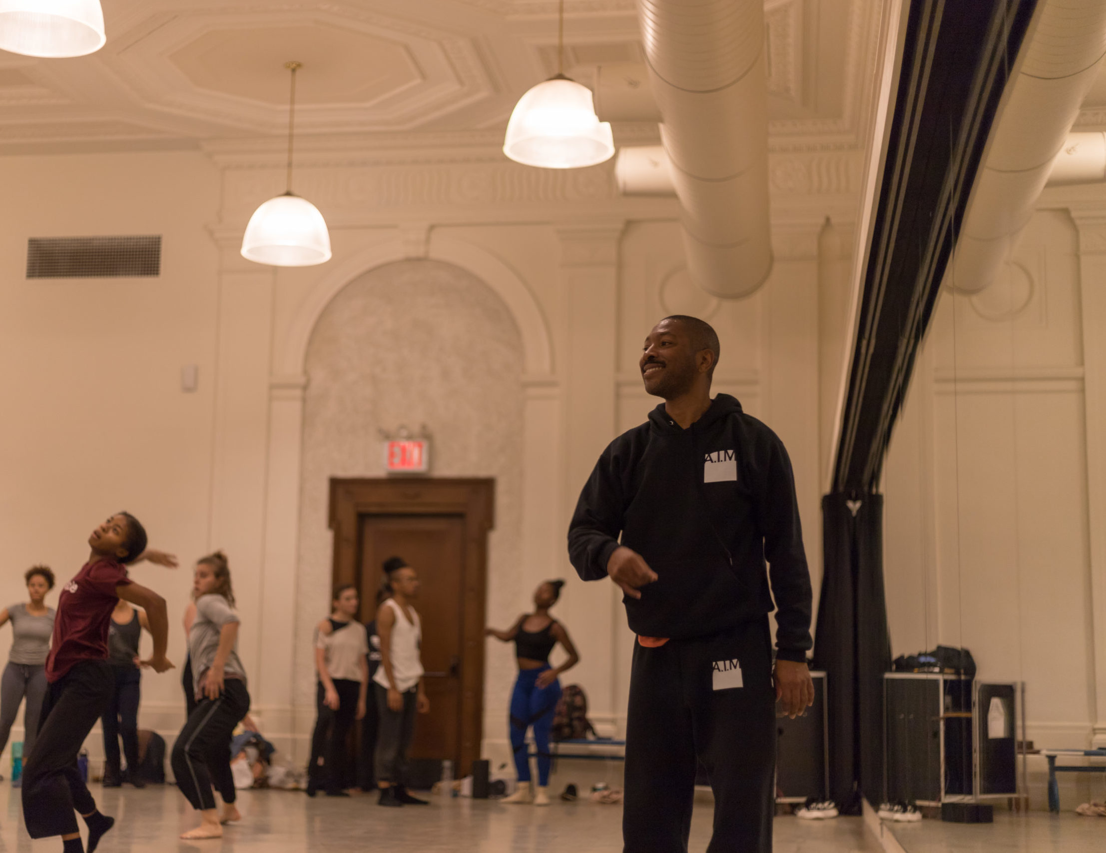 Kyle Abraham, wearing a hoodie with the A.I.M logo over the chest, smiles as he watches a group of dancers from the front of a large studio. One hand rises past his sternum as he goes to make a gesture.