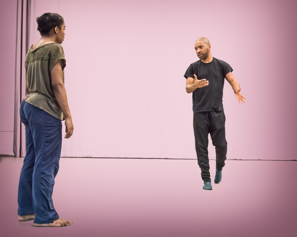 In a light purple–lit space, Kyle Abraham, dressed in black rehearsal clothes and socks, walks forward and gestures as he speaks to one of his dancers, listening with her back to the camera.