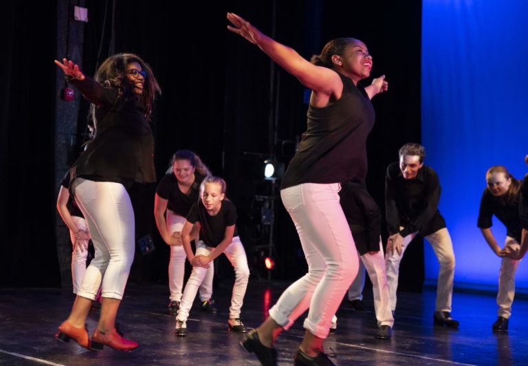 A group of teenage dancers performs onstage in white pants, black shirts and tap shoes