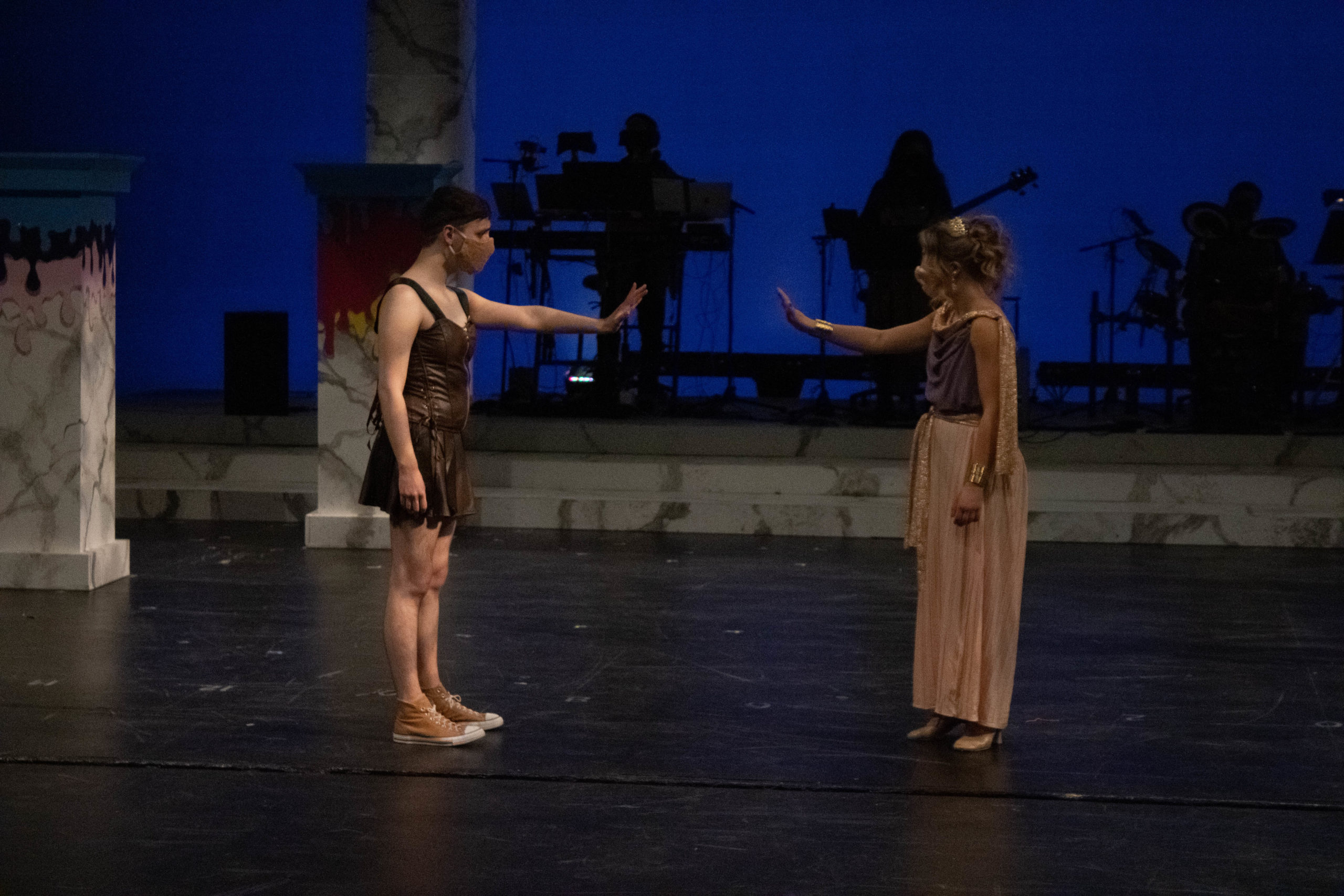 Two masked performers, a young man and a young woman, stand six feet apart onstage, reaching towards one another.