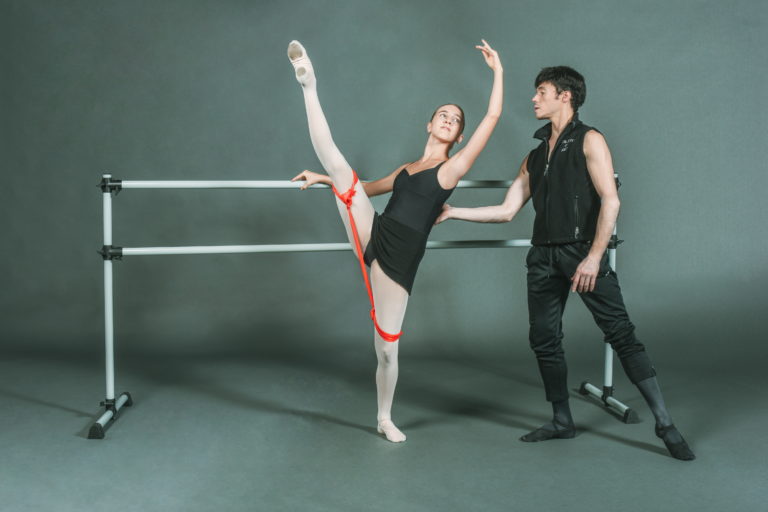 A teenage girl performs a battement at the barre, a Theraband tied to both her legs. Joseph Gatti stands behind her, adjusting her back placement.