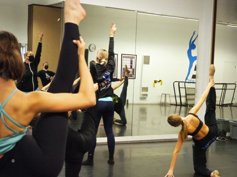 Sunny Reichert, seen from behind, stands at the front of the studio, pointing into the air. Her students behind her, some standing and some on the ground, extend one leg in the air.