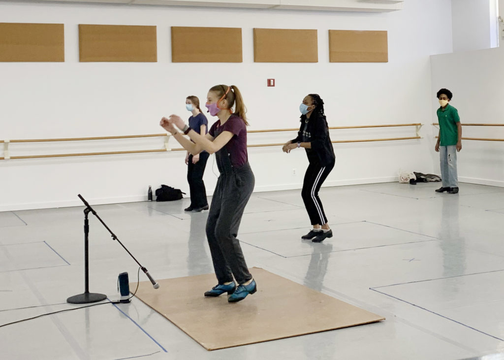 Three students of various ages take a tap class from a teacher who dancers on a tap board and has a microphone. Everyone is masked, and the students dance in boxes marked on the floor.