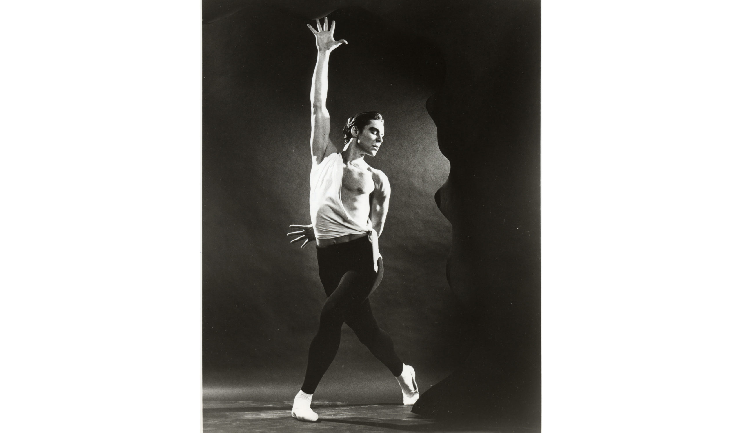 Black and white image of Jacques d'Amboise in black tights and a loosely-tied tunic showing half a bare chest, one hand splayed and raised to the ceiling and the other splayed and peeking out behind his lower back.